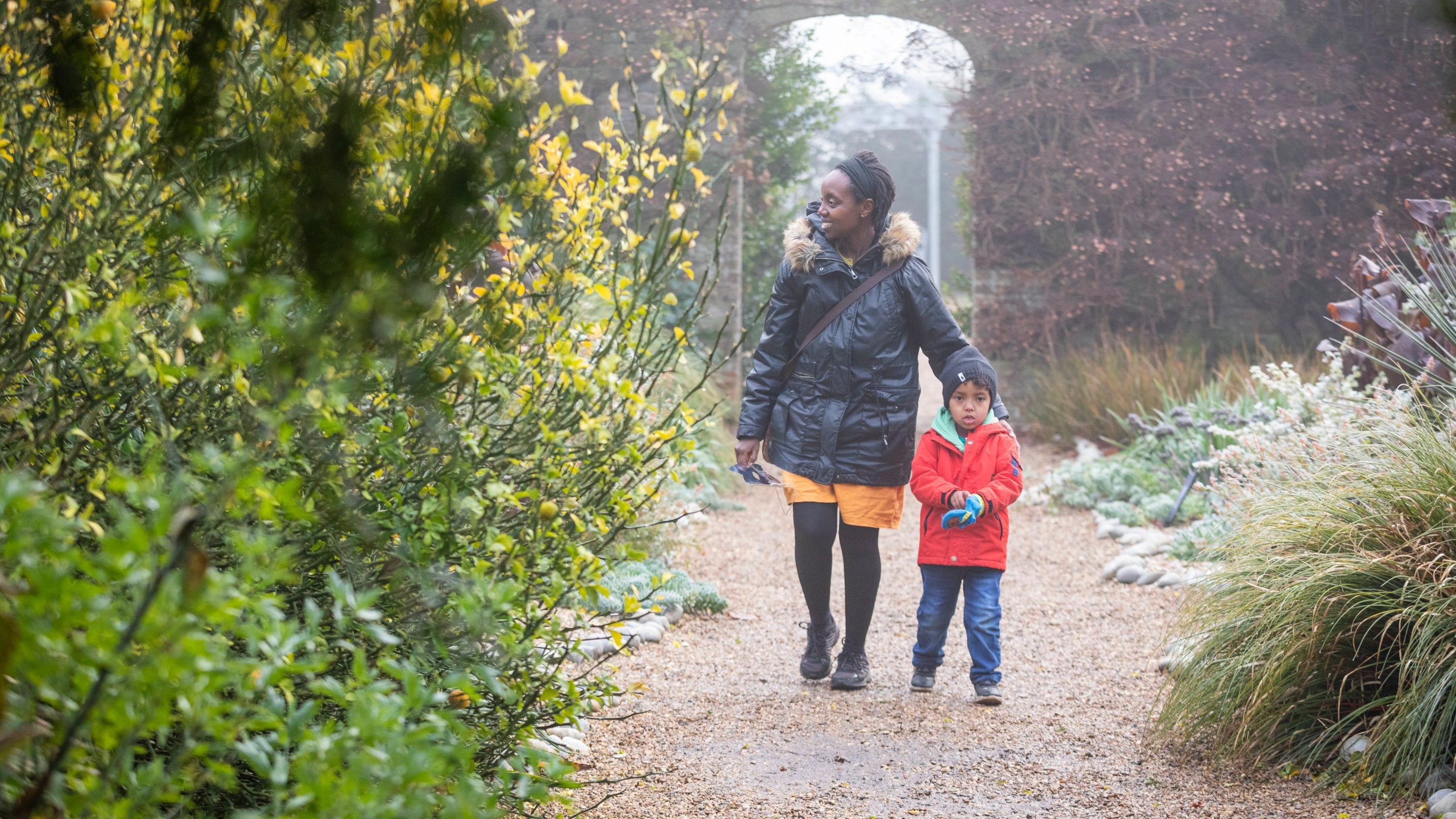 A woman and child walking along a path in a wintery garden with an arch in a red-brick wall behind them