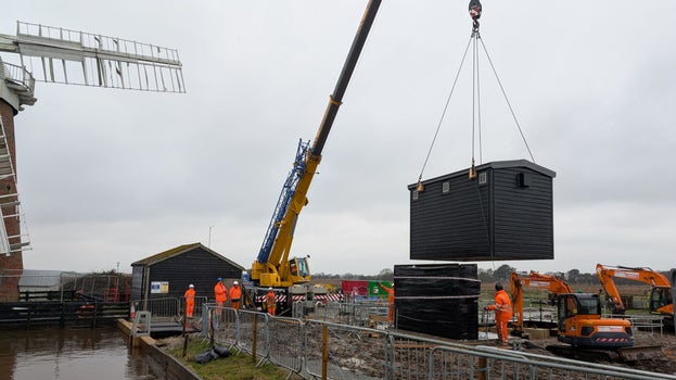 A crane lifts a shed like building into place above a rectangular metal structure that houses the pumping station controls