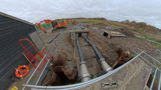Two exposed underground pipes can be seen running past the black wooden building that houses the existing pumping station, with construction vehicles in the background.