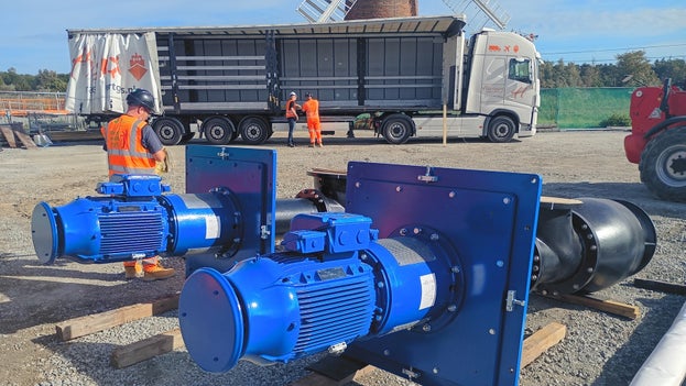 Two large pups lying on their side in the site compound with the lorry that delivered them in the background and Horsey Windpump visible further behind.