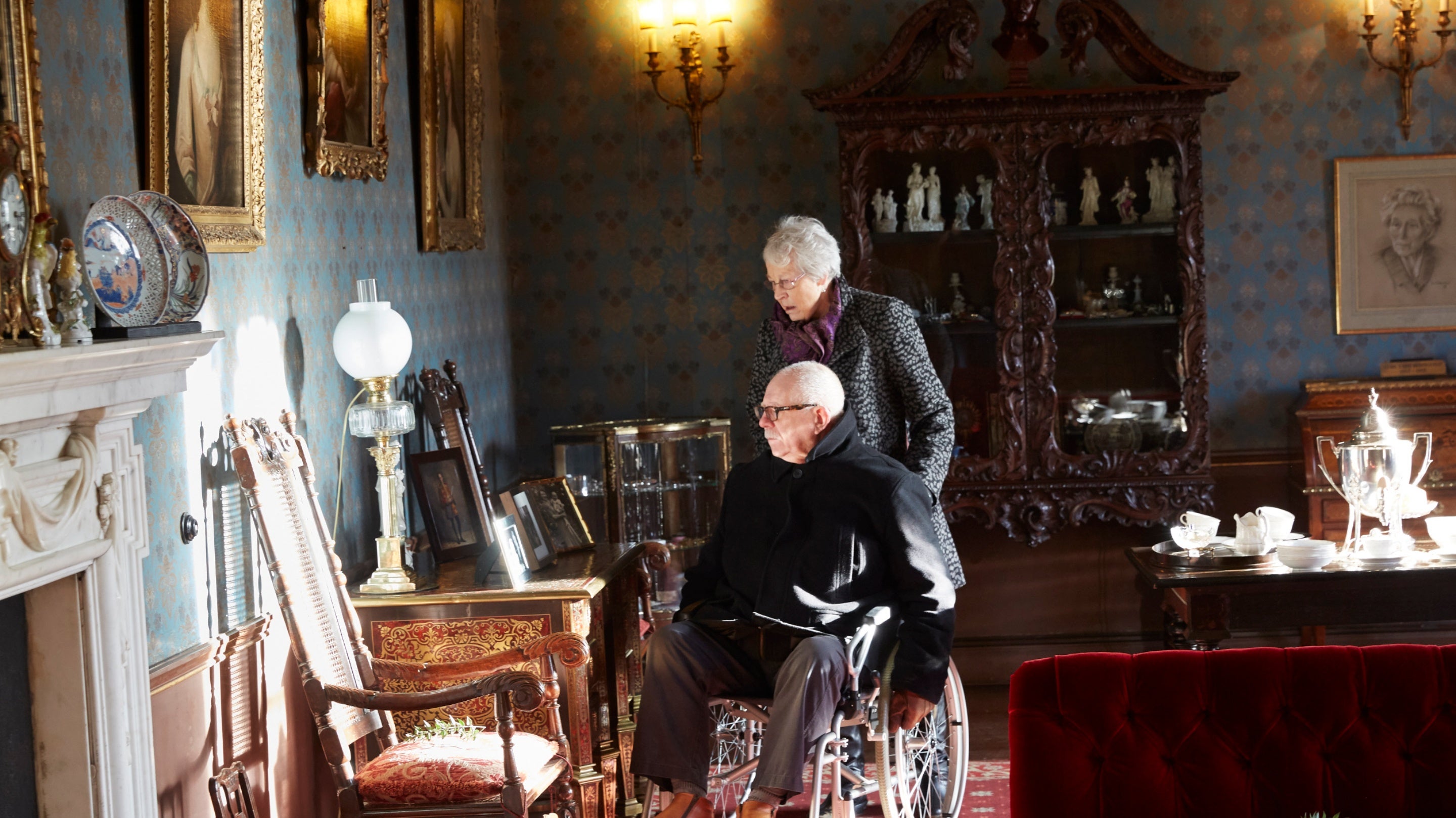 A lady pushes a man in a wheelchair as they look at photographs on a table in a room with lots of period collection items