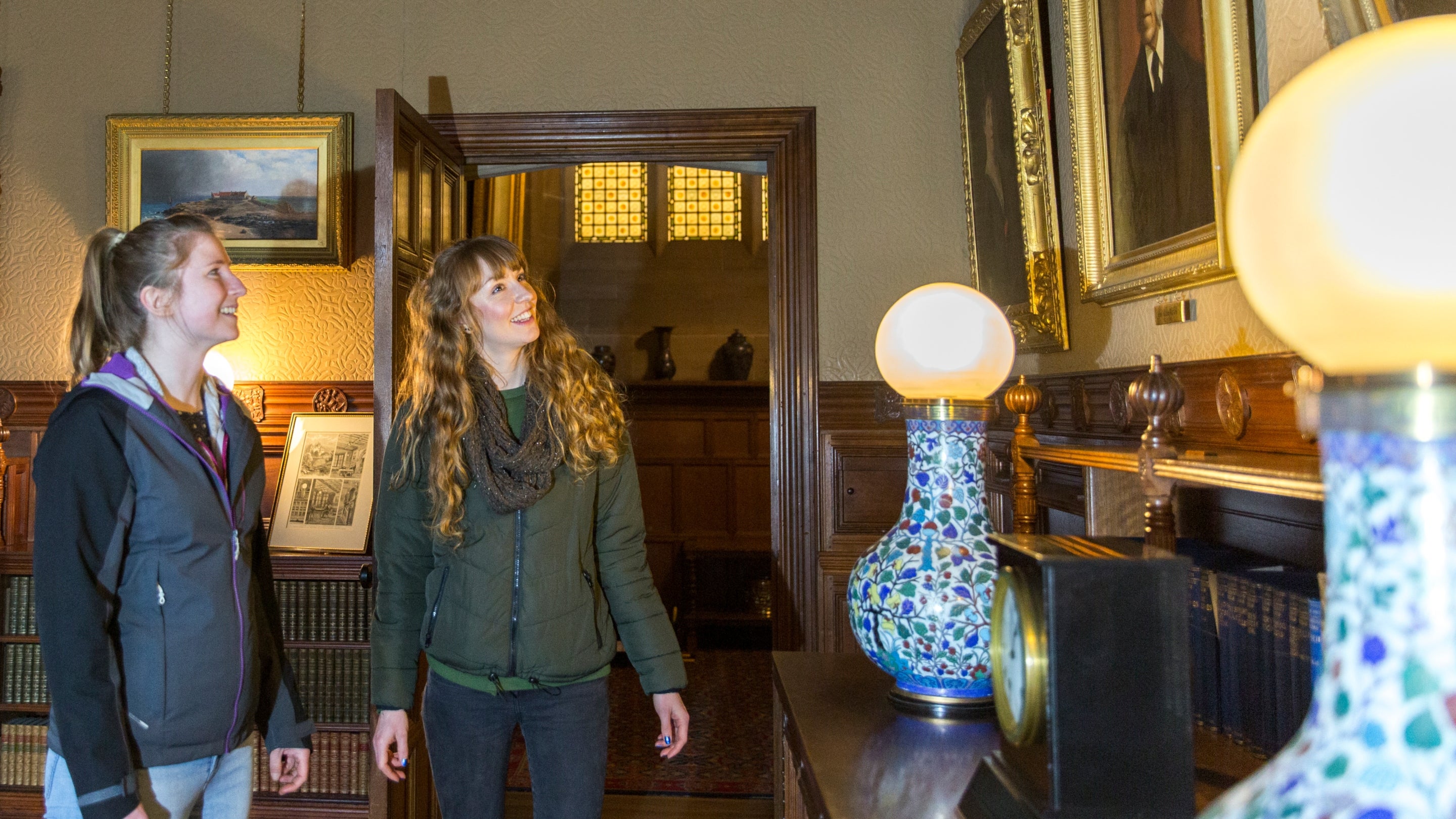Two visitors in the Library at Cragside. Two lamps lamps are the focus on the side of the image, they are round glowing bulbs on the top of ornate Chinese vases.
