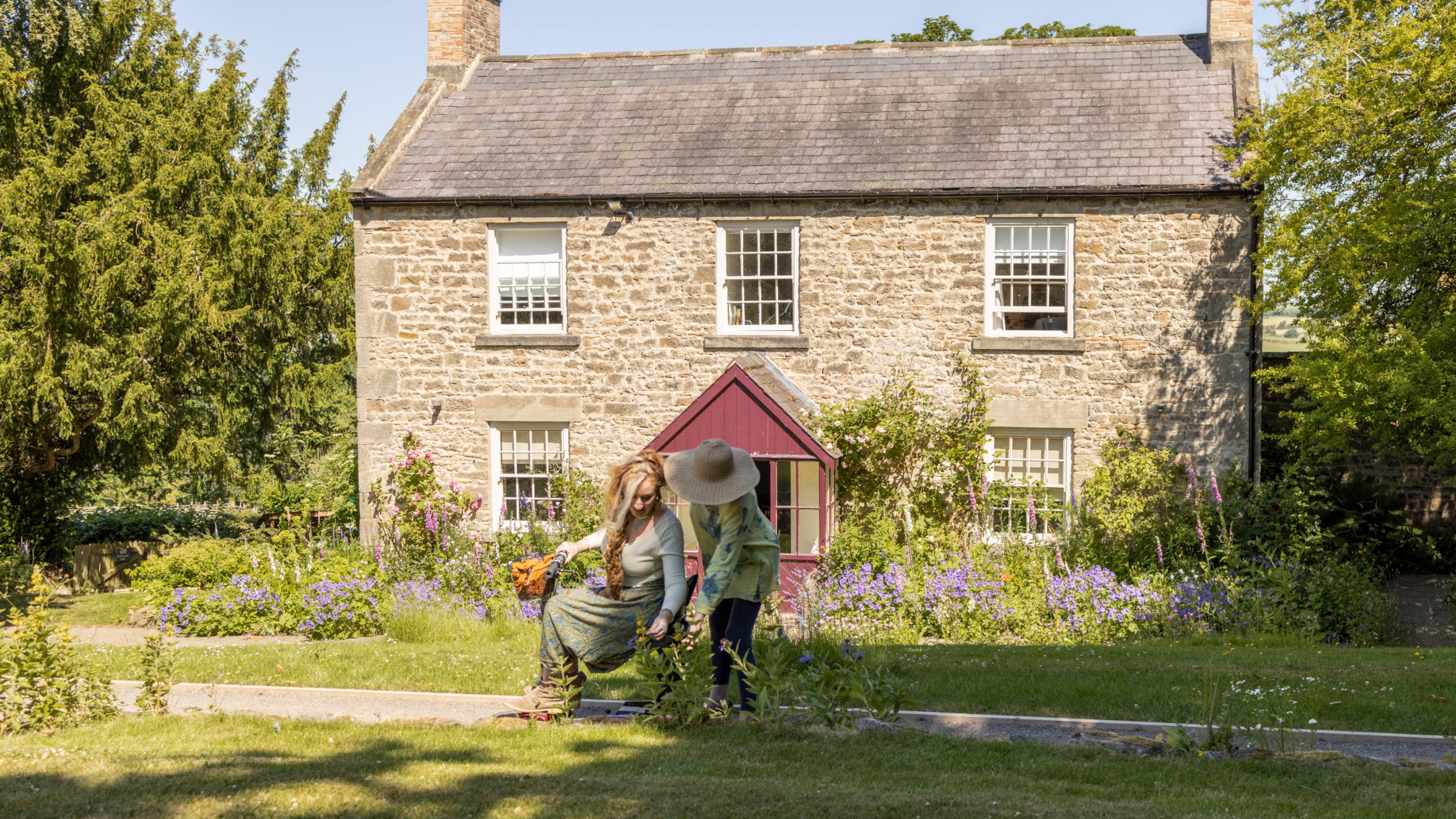 Two people, one a wheelchair user, admiring flowers in front of a stone-built house