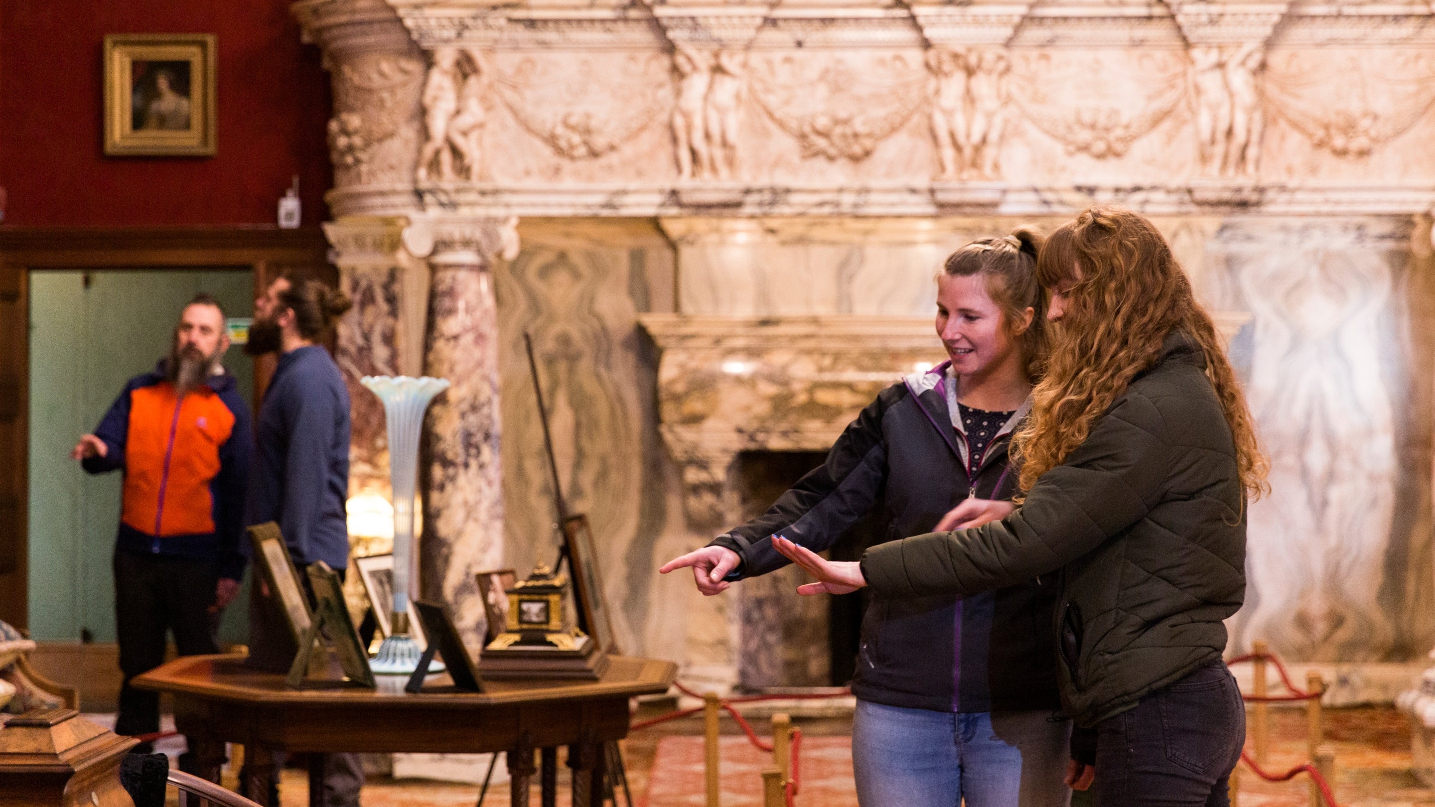 Part of a group are in the Drawing Room at Cragside House. The room is decorated in rich red tones and a grand, white, decorative, marble fireplace is in the background. Two visitors are chatting and pointing to an object on display.