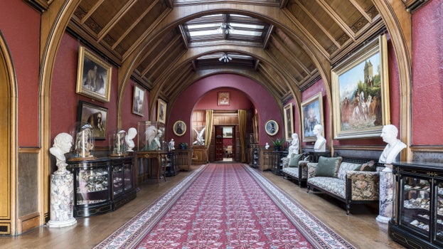 Art lines the dark pink walls of Cragside's Gallery. The ceiling is arched with a skylight and the rooms is dressed with dark wooden cabinets and long seating