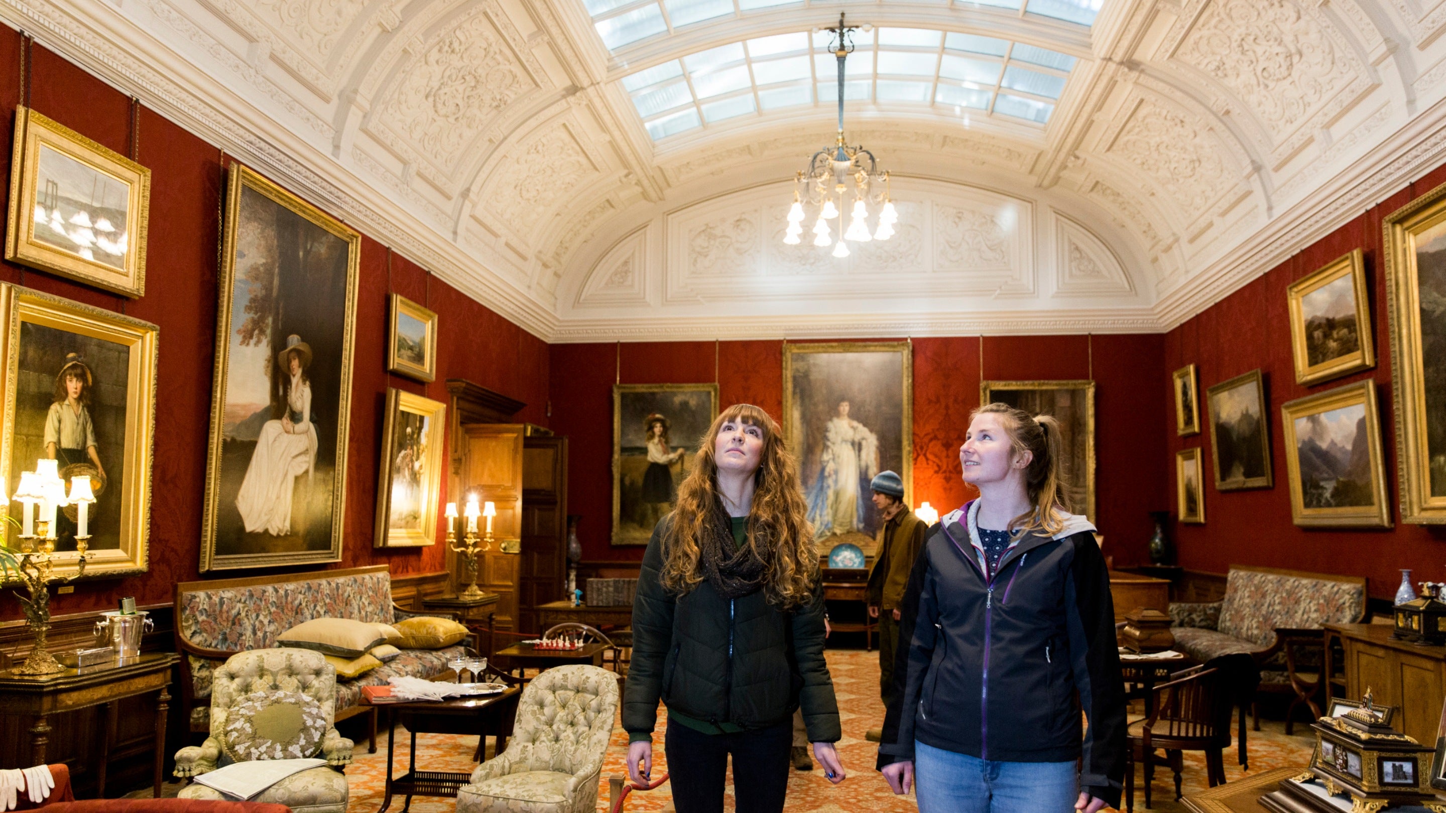 Two visitors in a room with a white vaulted ceiling with a sky light with dark red walls and various artworks displayed.