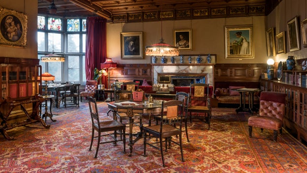 The Library at Cragside House with luxurious red furnishings including the velvet curtains, upholstered furniture, plush carpet and hanging lampshades.