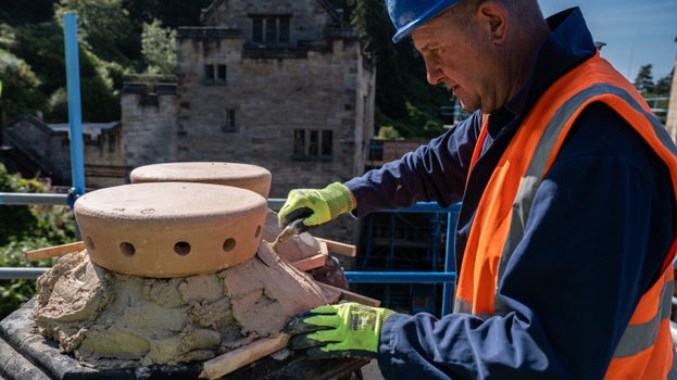 A contractor wearing a high-visibility vest and safety helmet repairs stonework on a rooftop at Cragside. The contractor applies mortar around a newly installed sandstone chimney pot, with the house visible in the background.