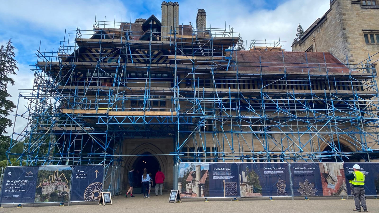 Cragside House covered by a large free-standing scaffold structure during conservation work, with visitors entering through an archway beneath the scaffold and interpretation banners lining the front.