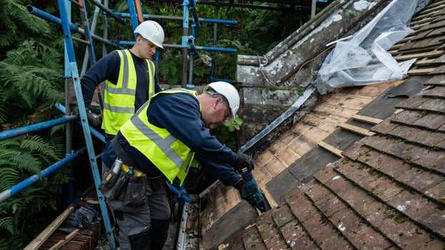 Two contractors wearing high-visibility vests and hard hats repair a section of Cragside's roof. Roof tiles have been removed, exposing the timber structure beneath, while scaffolding surrounds the work area.