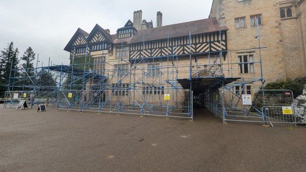 Cragside House partially covered in blue metal scaffolding. Temporary fencing and warning signs surround the structure. The sky is overcast and the ground appears damp.