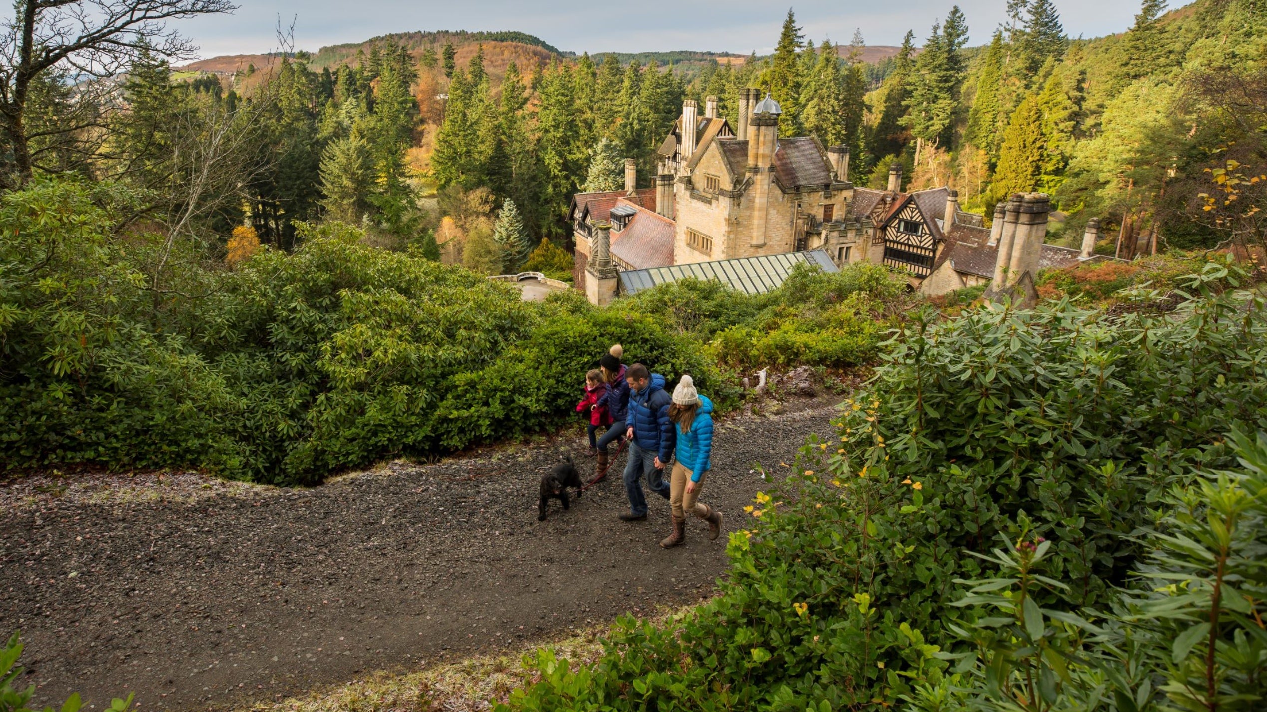 A family, 3 adults, 1 child and a black dog, are walking along a path at Cragside. The chimneys of Cragside House are visible. The day is cold and wintry. Everyone is wearing bobble hats.