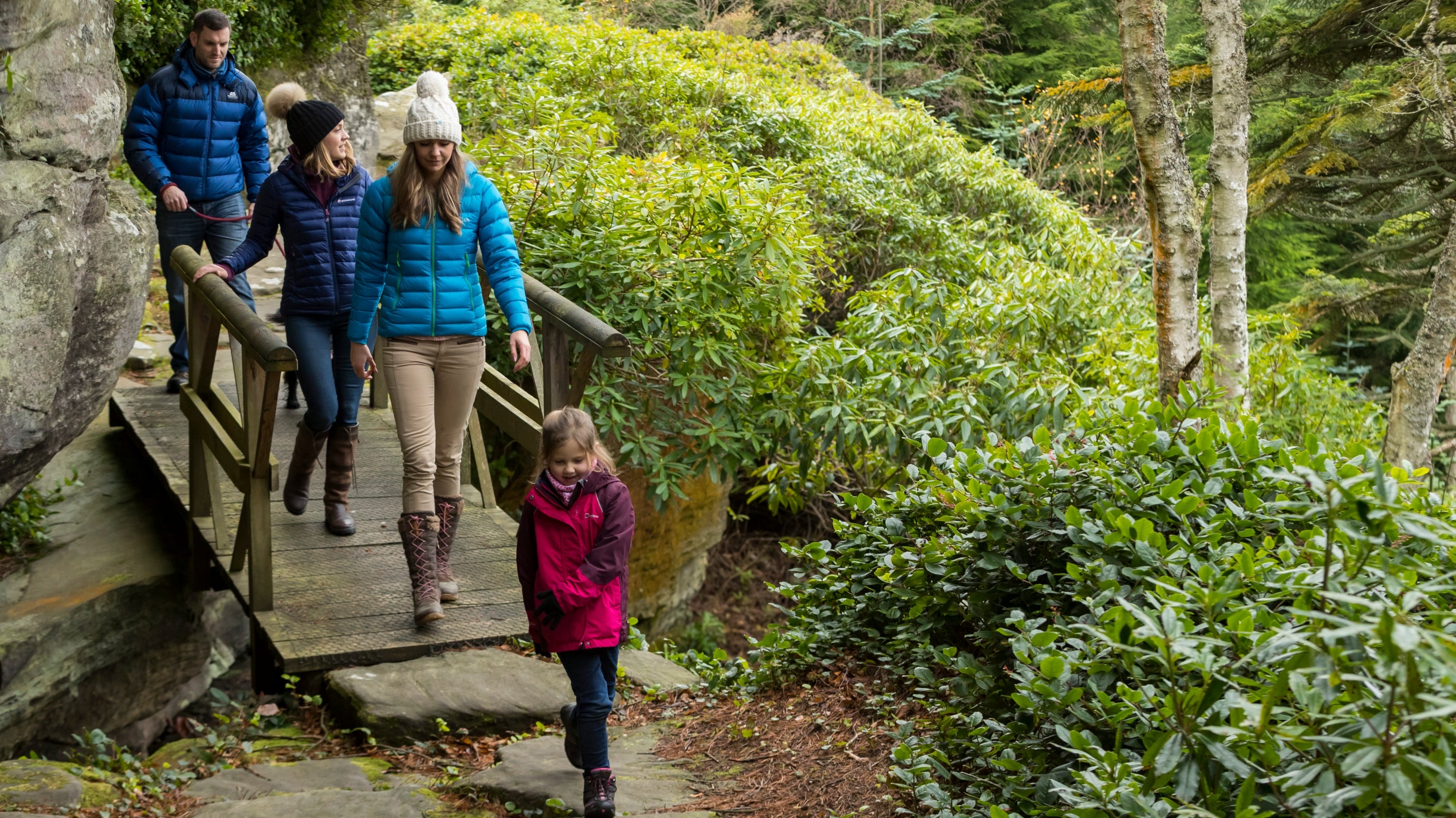 A group of four people in outdoor winter clothing walk across a small wooden bridge on a woodland path surrounded by rocks, shrubs and evergreen trees. A child in a bright pink jacket leads the way along the path, with three adults following behind through the lush green landscape.
