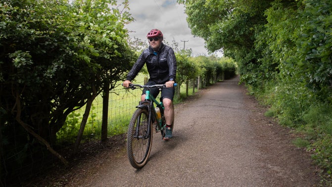 A cloudy but bright summer's day. A man on a mountain bike in cycling gear and helmet rides towards the camera along a country lane with a fence and gappy hedge on one side, dense trees on the other.