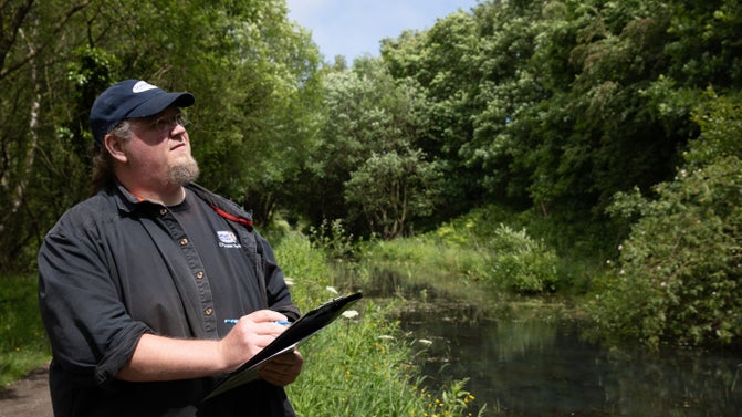 A ranger in a black uniform and cap with a Durham County Council logo holding a clipboard and pen, with lush vegetation, trees and a pond in the background.
