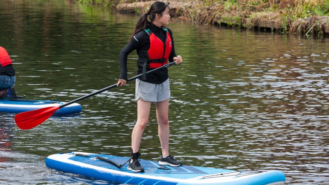 A stand up paddle boarder standing on a blue paddle board, wearing a red life jacket and holding a red paddle moves along a wide river. Behind is the end of another paddle board and a grassy river bank.