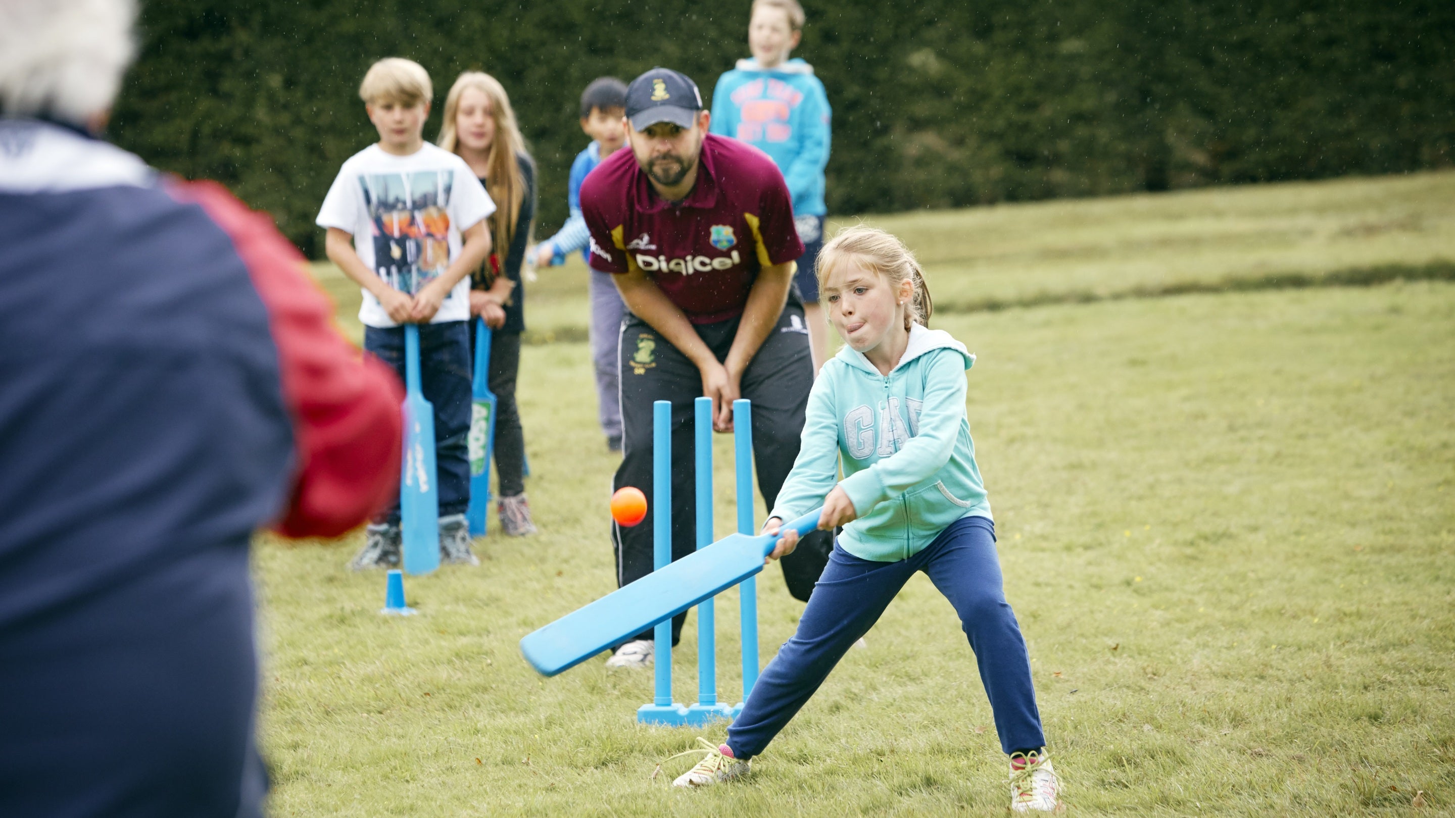 Children play cricket on a grassy field in summer. Adults in cricket club kit are bowling and fielding. One child bats, while others line up to take a turn.