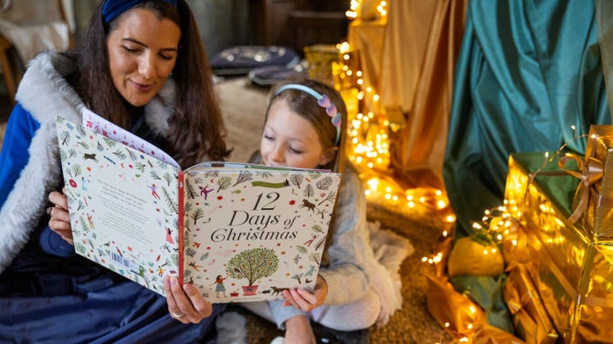 A young girl and a woman in a blue cape read 'The 12 Days of Christmas' in front of fairy lights.