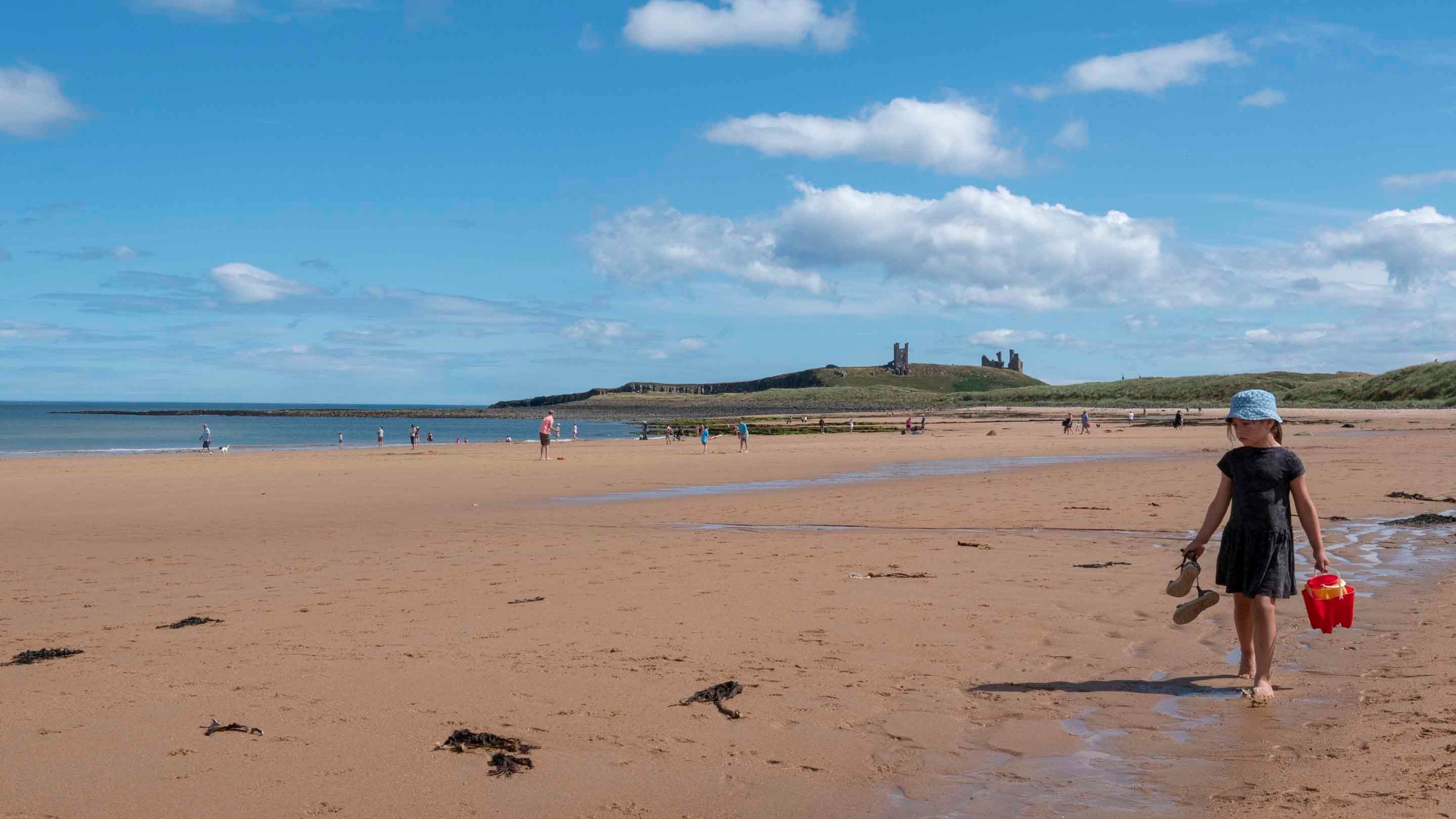A girl in blue hat with a red bucket walks along a sandy beach with Dunstanburgh Castle visible on a hill in the background.