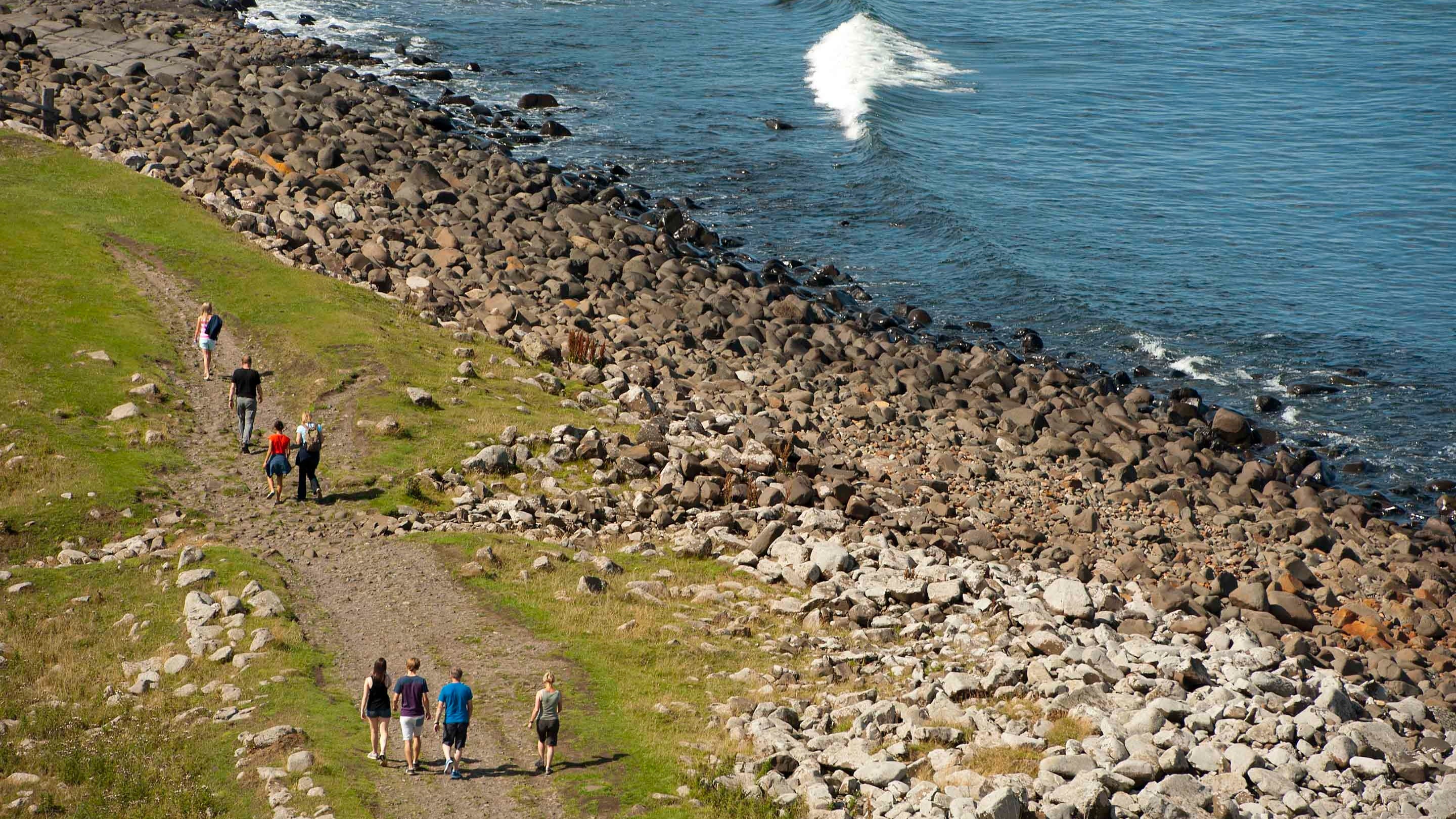Aerial view of people walking along a rocky path by the coast, with crashing waves, at Dunstanburgh Castle