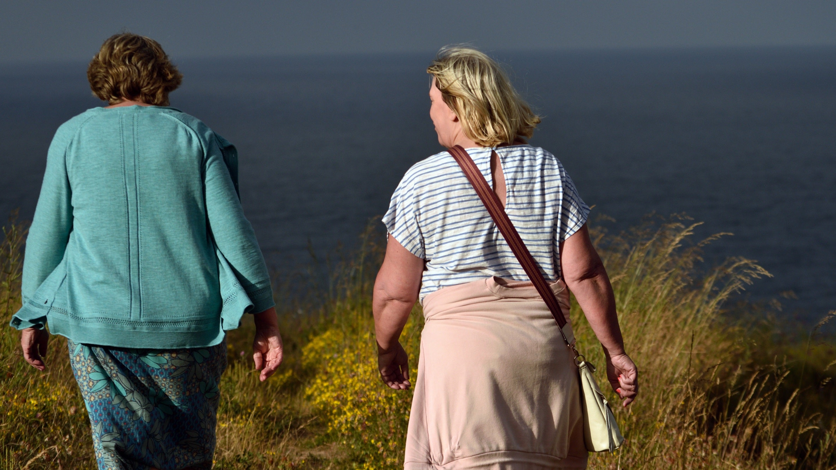 Close up of two walkers on a grassy clifftop in low sunlight with dark blue sea beyond