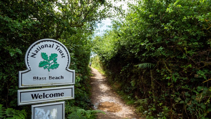 View of a path through hedgerows. To the left is a National Trust sign for 'Blast Beach'.