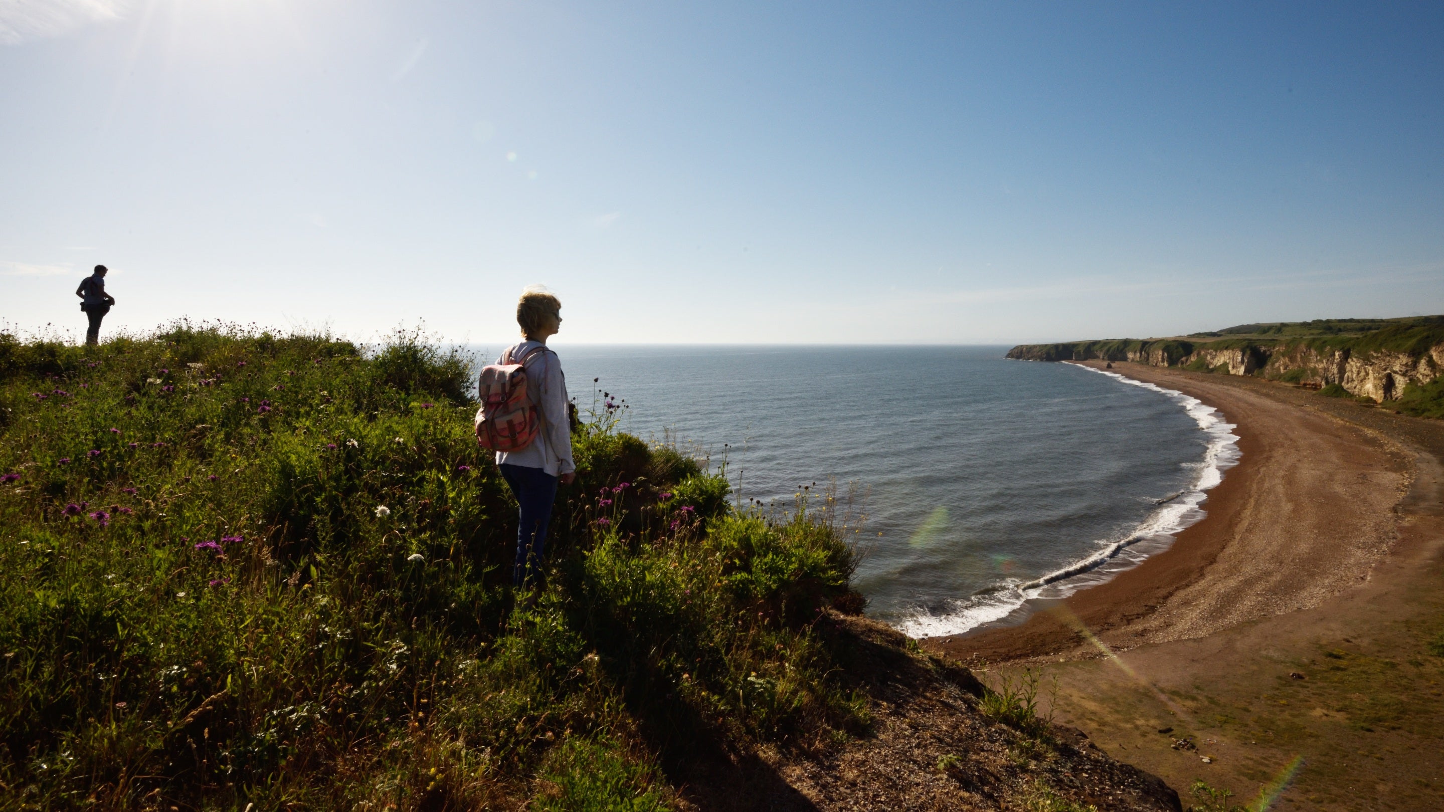 Two people, one silhouetted against the sky, stand on a grassy cliff top in the morning sun. They're looking over the curved bay and pale rugged cliffs of Blast Beach near Seaham.