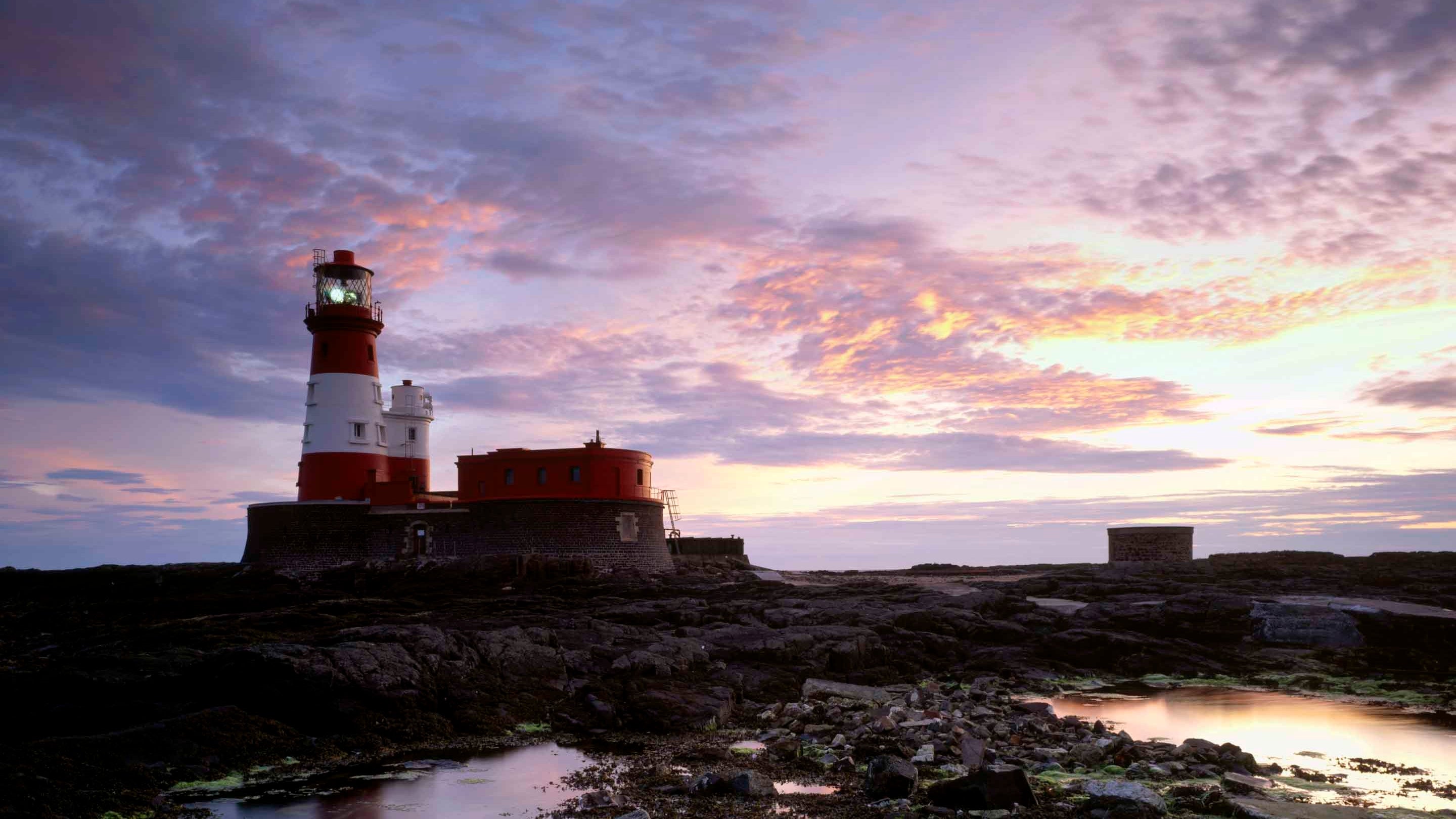 A sunset view of Longstone Lighthouse across a rocky area with pools of water on the Farne Islands, Northumberland