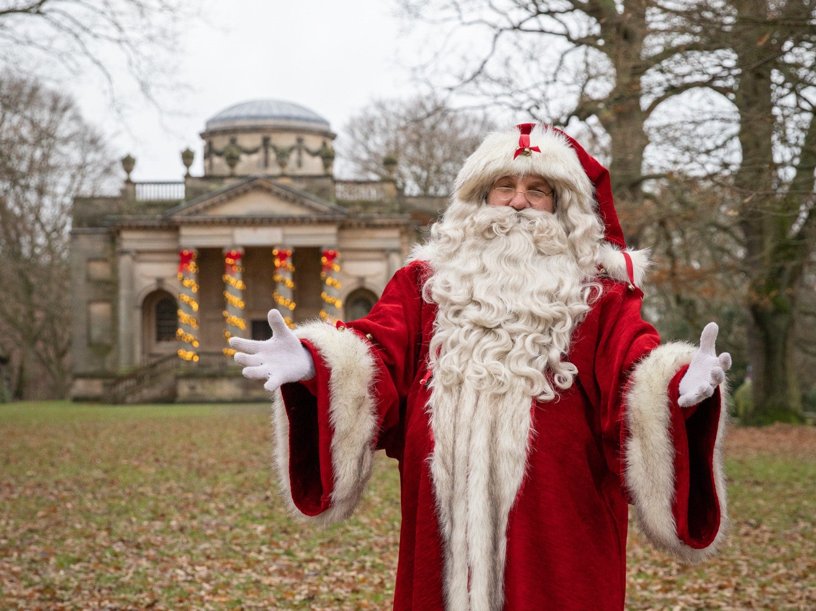 Father Christmas in traditional red clothes and a big white beard in front of a Chapel covered in Christmas lights.
