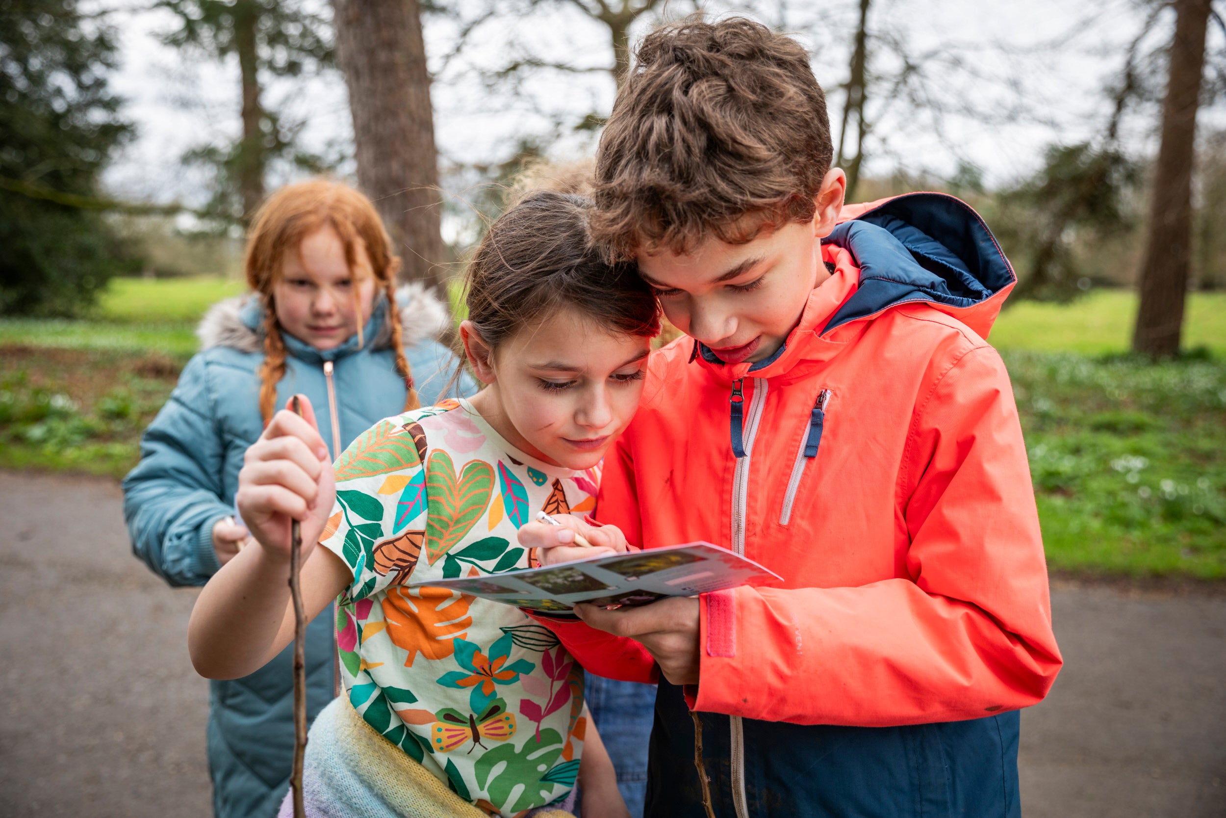 Two children looking closely at a trail sheet, with another child and greenery in the background.