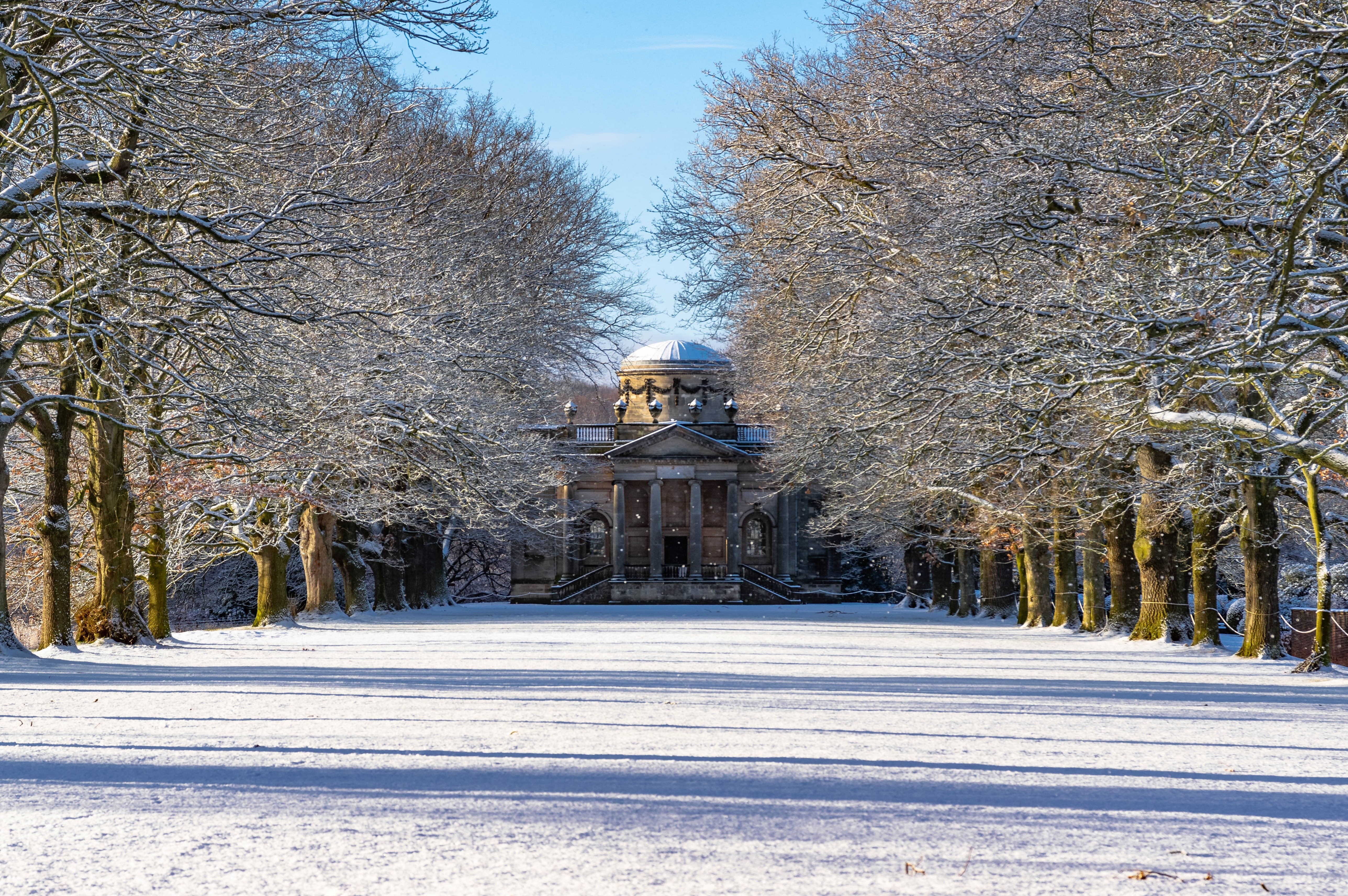View of a snowy avenue of bare trees leading to a columned chapel covered in snow.