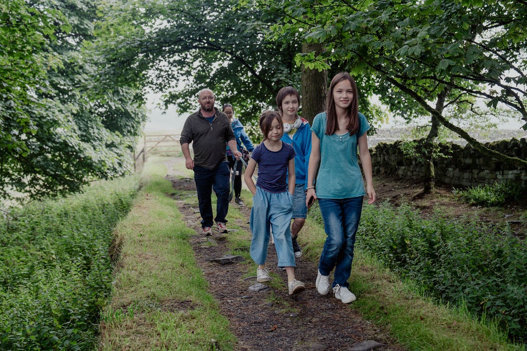 A family group of three children and two adults are walking along Hadrian's Wall. There are trees in full green leaf either side of them forming a canopy above.