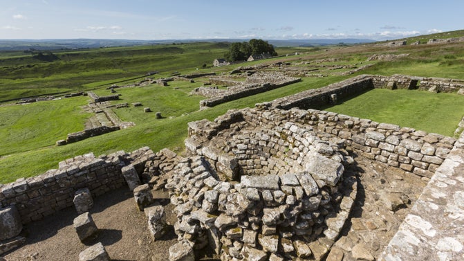 A view over grassland with low-lying ruins in the middle distance and foreground with blue skies above