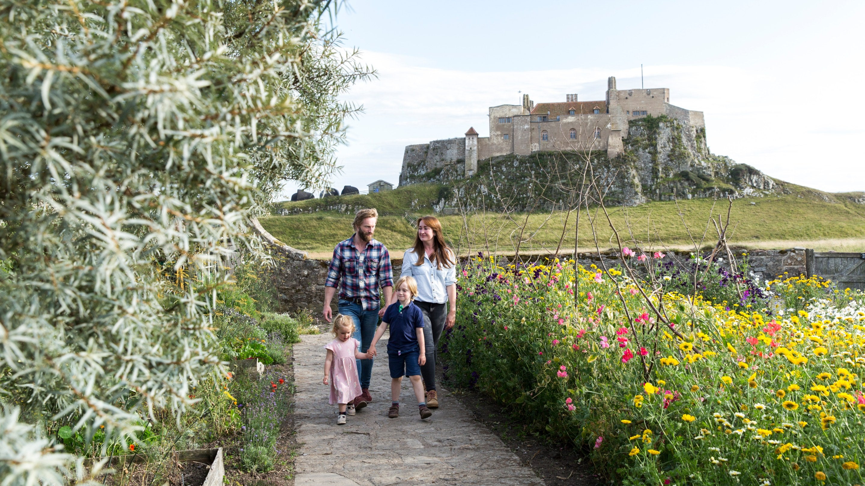 A family walk down a path with a summer garden to the right and a castle in the distance beyond the garden.