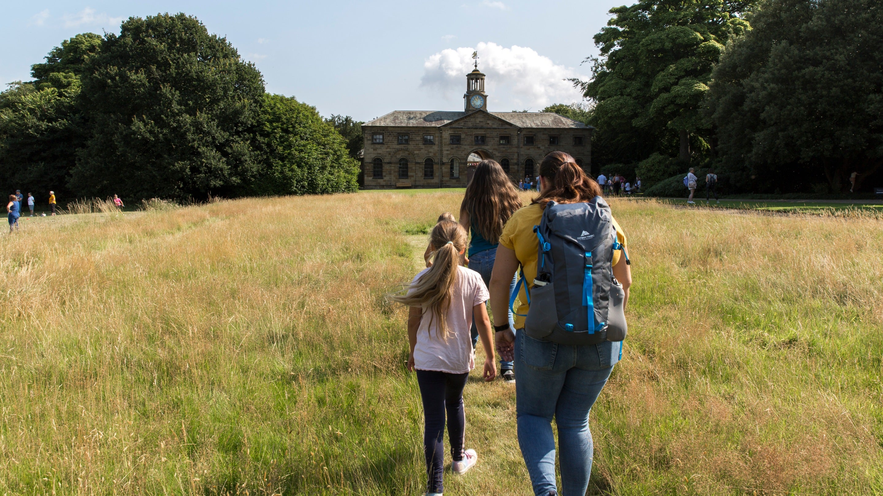 A mum and two children walk away from the camera towards the stables through the parkland along a mown path