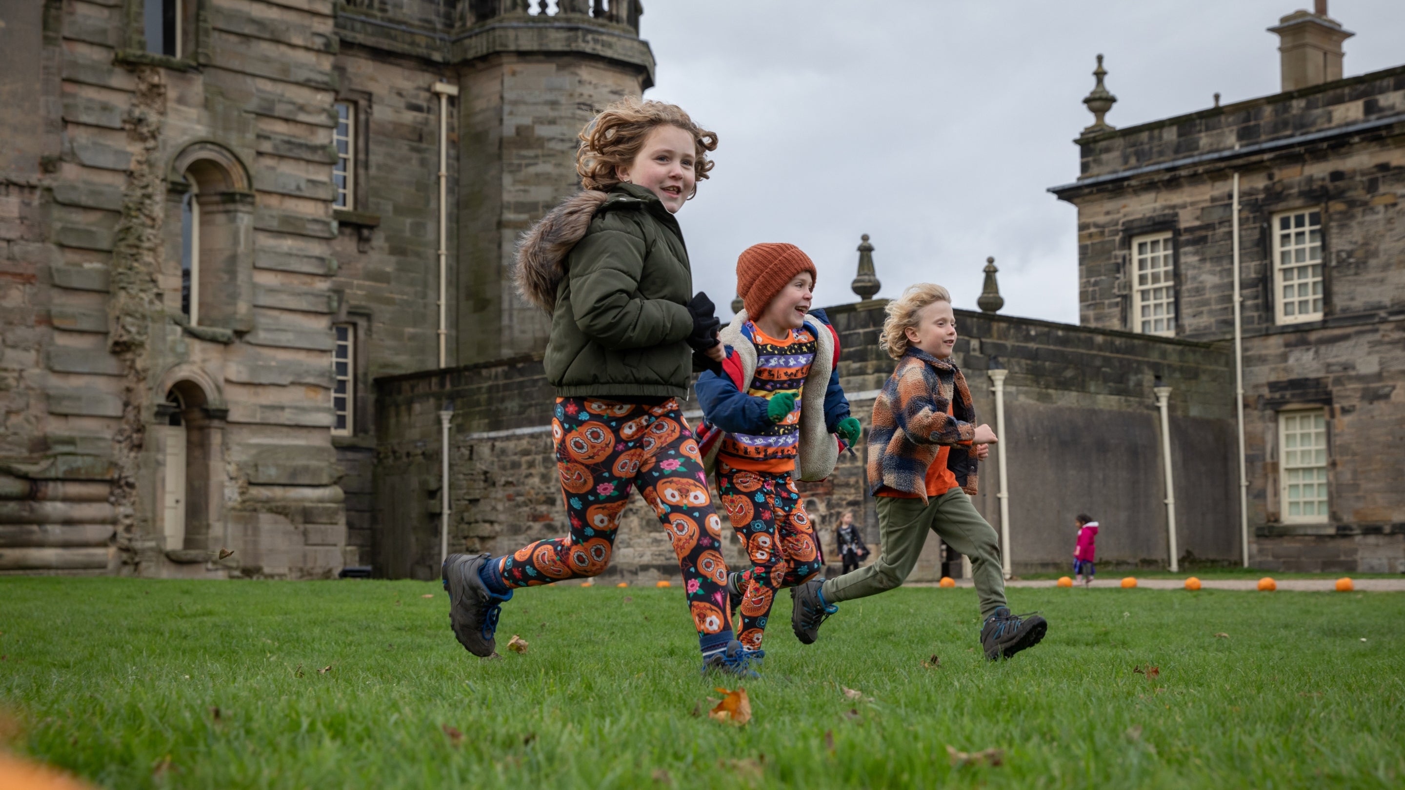 Three children running across the lawn in front of a grand Georgian hall on an autumn day, brown leaves and pumpkins are around on the grass.