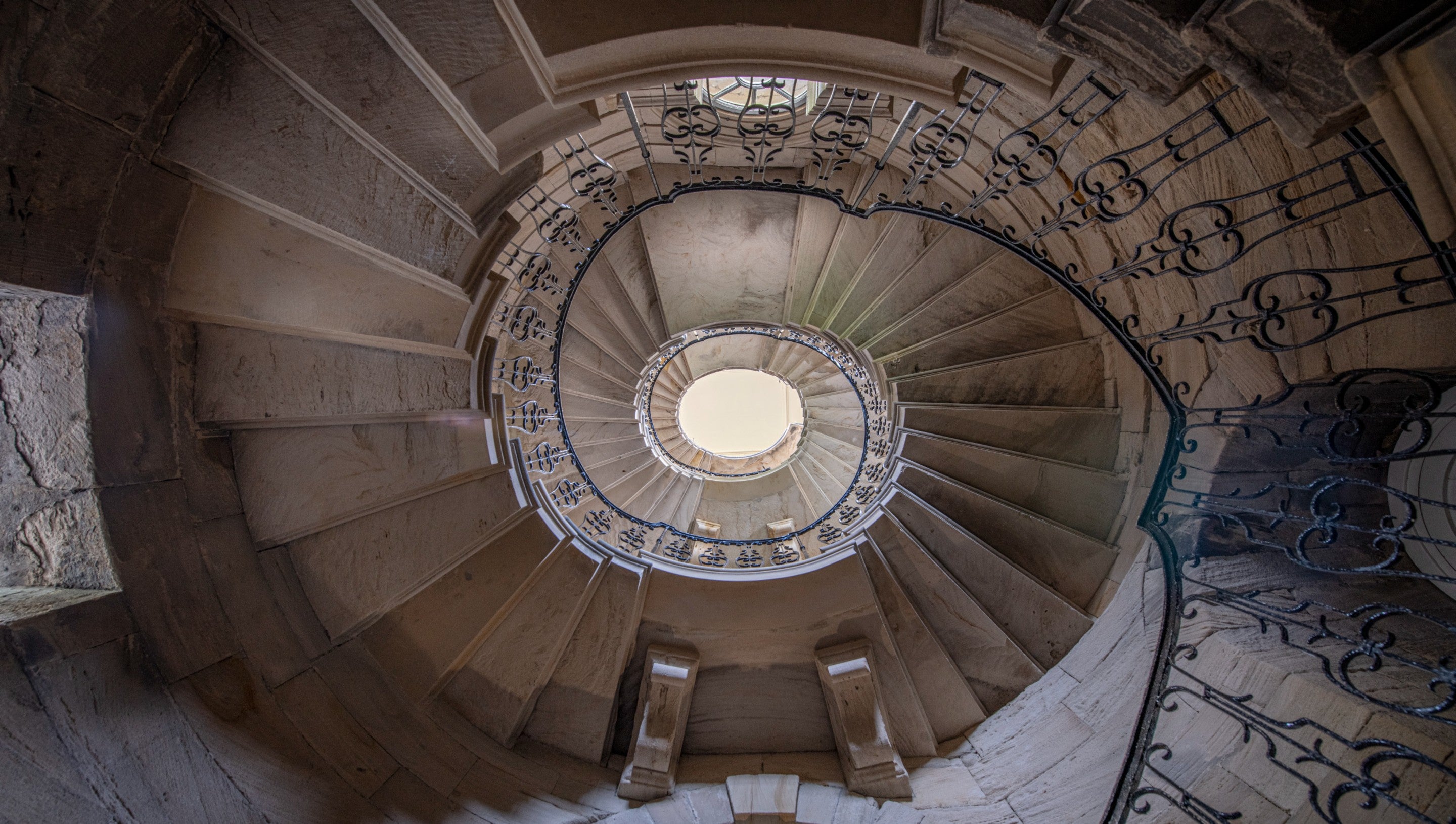 An upward perspective of a set of spiral stone stairs. They appear as a swirling pattern and there are black painted iron railing surrounding them.