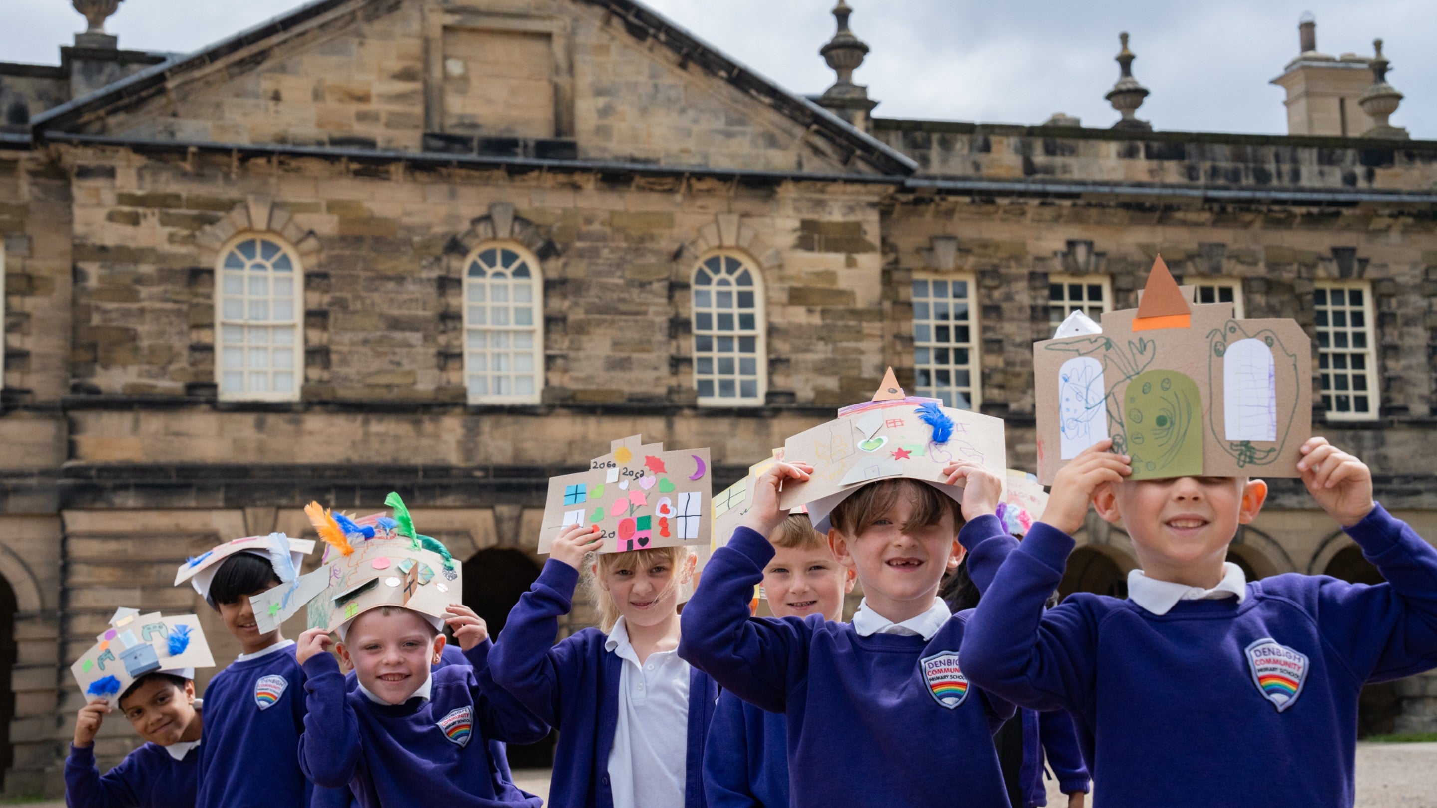 A group of schoolchildren standing together wearing cardboard hats in front of a stone building