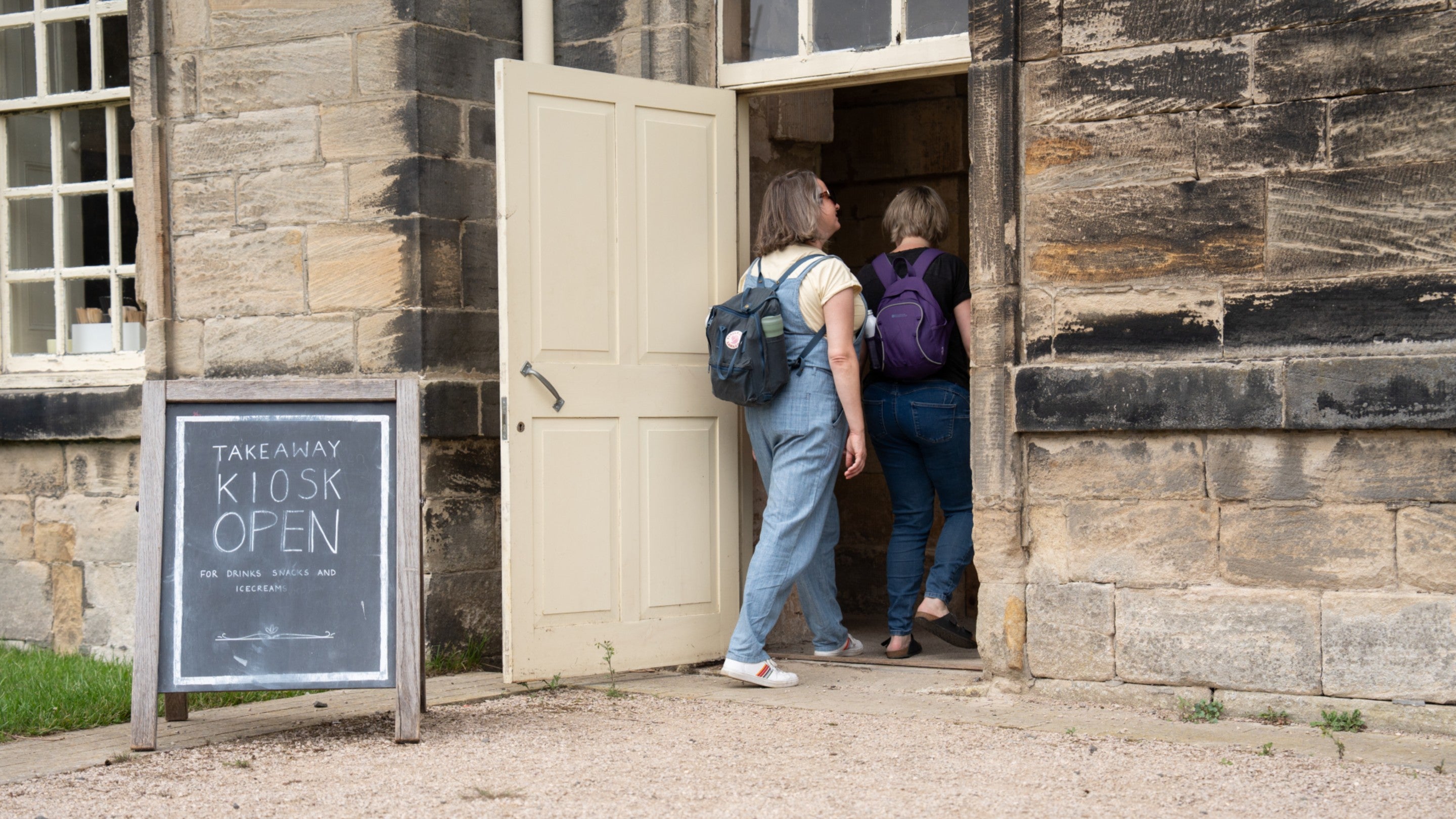 Two adult women are walking through a doorway. In front of the doorway is a sandwich board with the words 'Take away kiosk open' chalked on it.