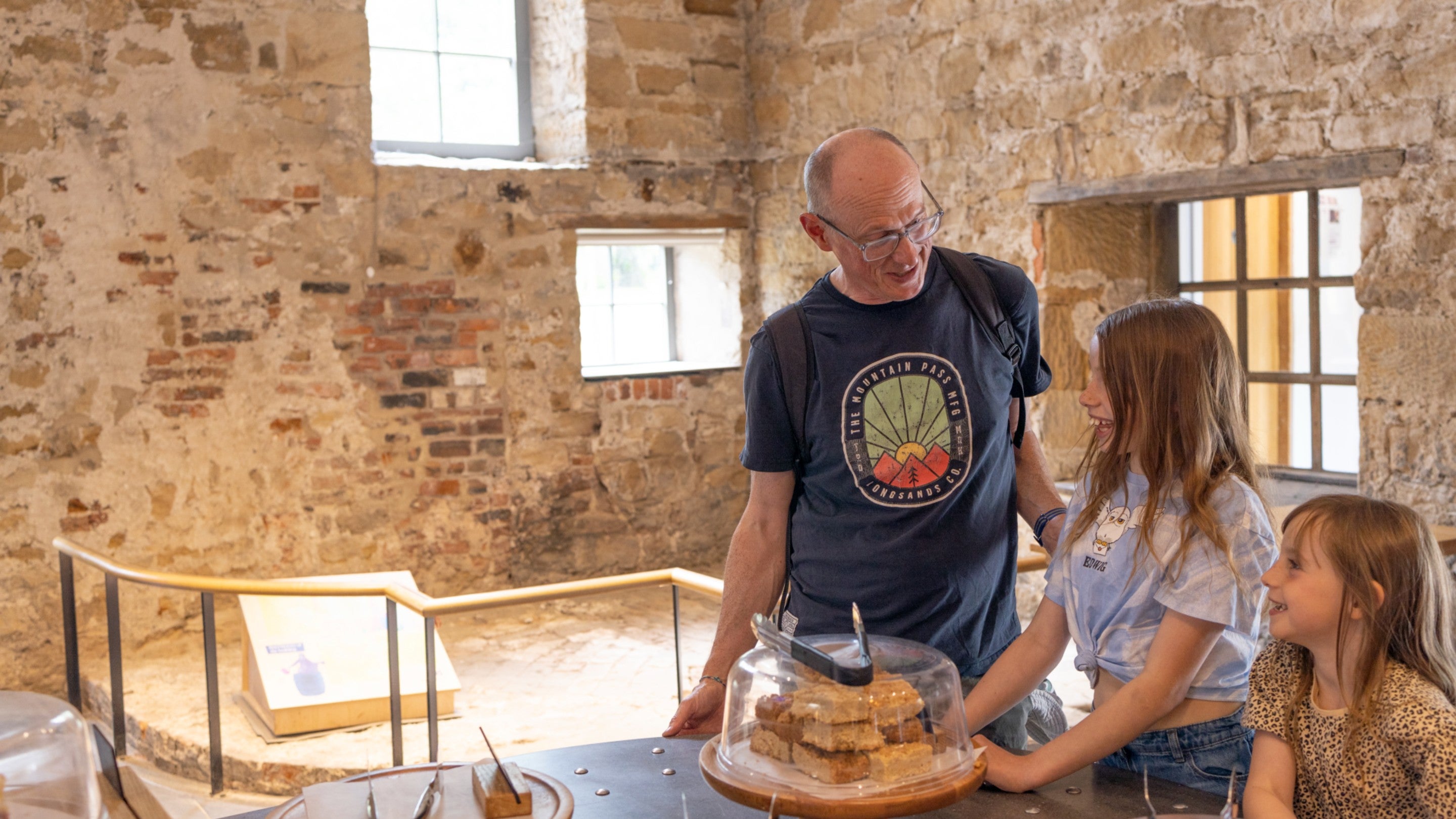 An adult white male and two white female children are standing inside a stone building with sunlight streaming through the windows. They are at a counter, on which is a tray of what look like flapjacks, which is covered with a glass dome.