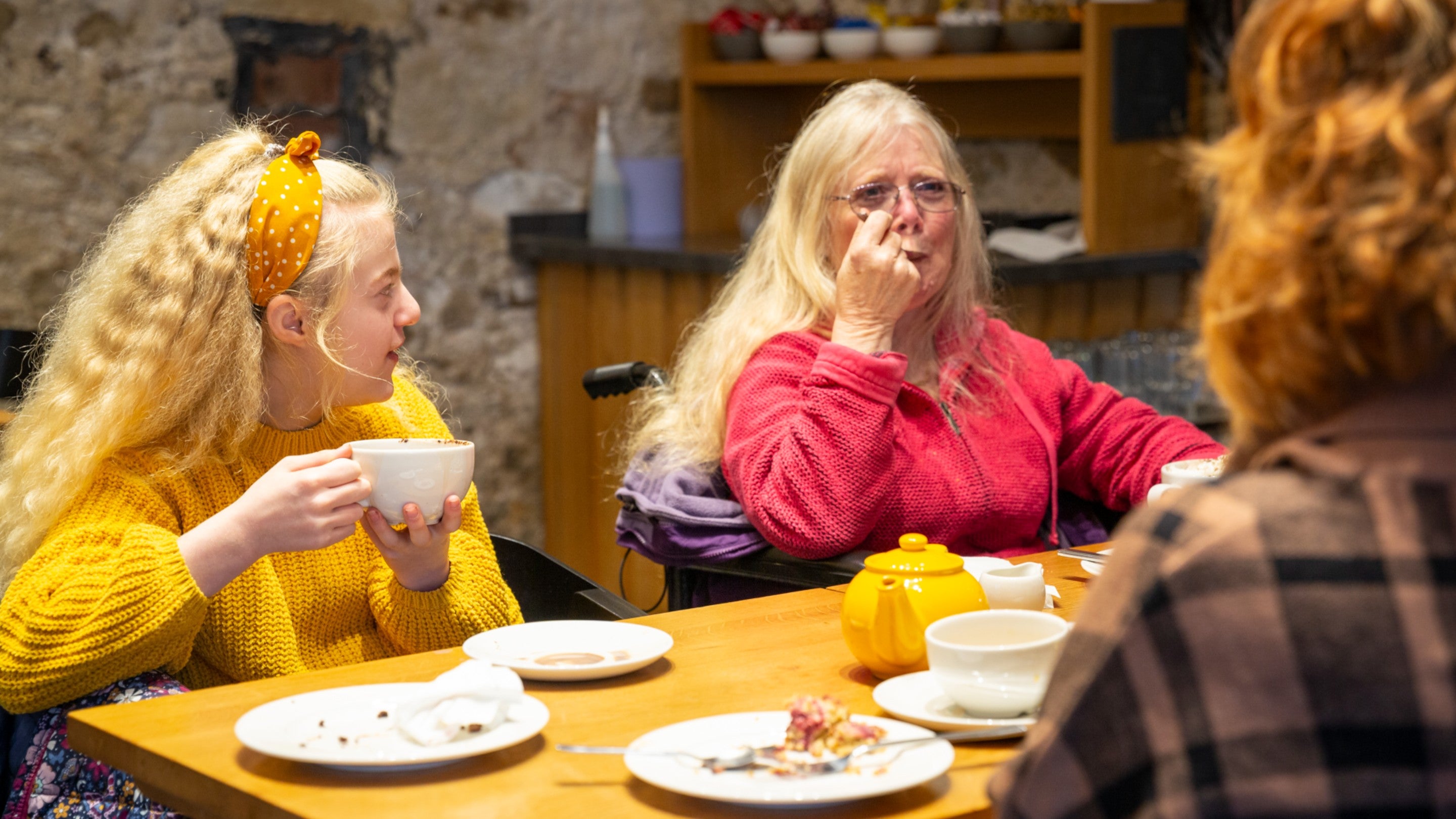A young girl with long blonde hair in a yellow jumper is sitting at a table holding a mug of hot chocolate. Next to her is an older lady in a pink jumper with long blonde hair and glasses. On the table in front of them both is a yellow teapot and several plates of cakes and scones.