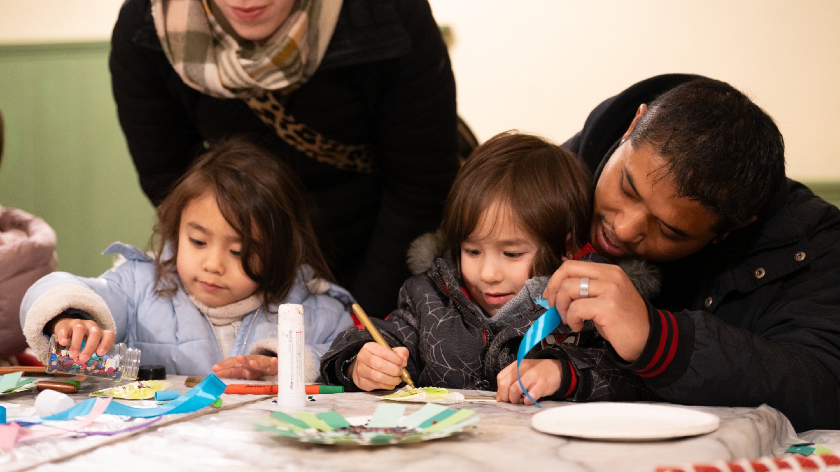Two young female children are sitting at a table. In front of them are crafting materials including paper plates, glue sticks and coloured strips of paper