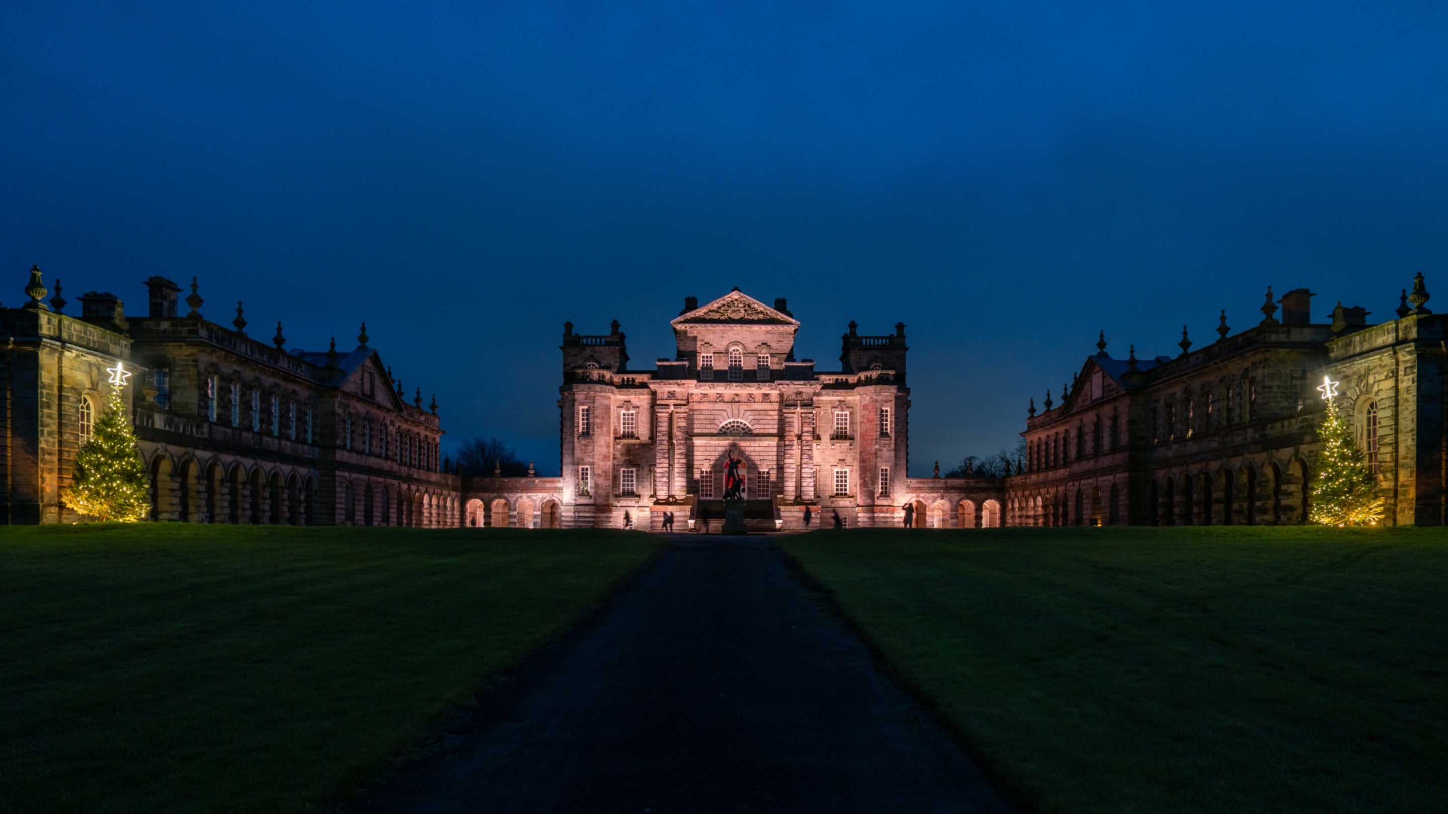 A large stone building flanked by two wings on each side. The central building is lit up in a pale pink colour. Behind the building the sky is a deep navy blue. In the foreground there is a central driveway with grass either side.