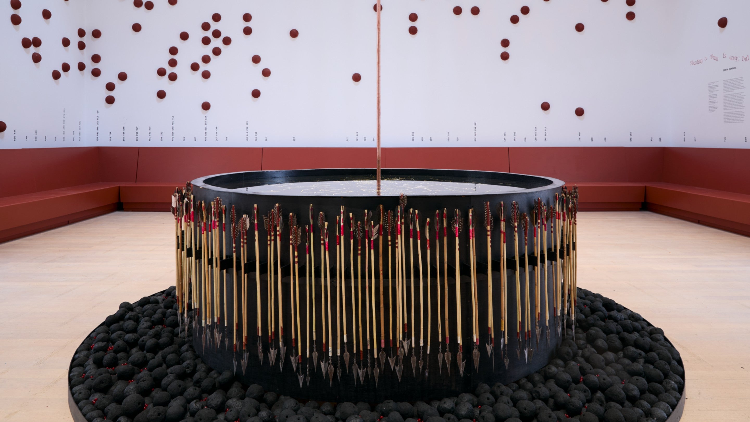 A sculpture with what looks like a large, black drum surrounded by spears pointing downward. At the base of the drum there is a cylinder of pieces of coal. The work sits inside an otherwise empty room.