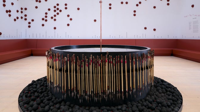 A sculpture with what looks like a large, black drum surrounded by spears pointing downward. At the base of the drum there is a cylinder of pieces of coal. The work sits inside an otherwise empty room.