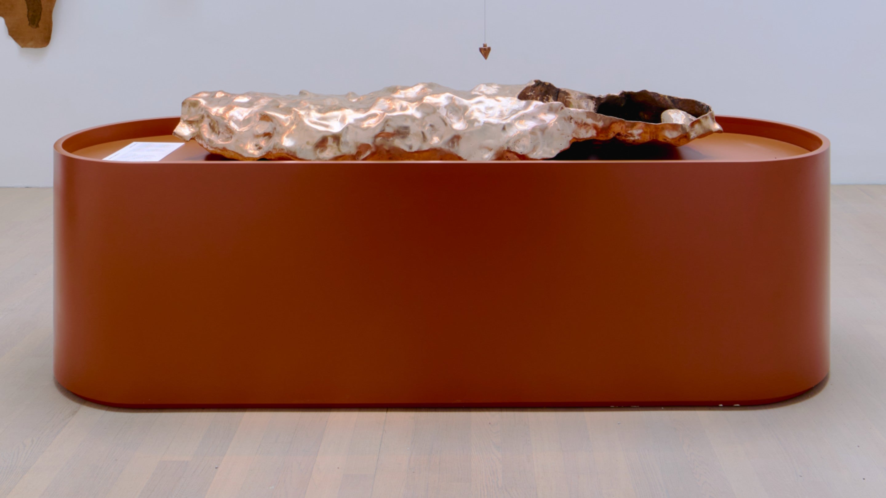 A metal sculpture sits centred on top of a cylindrical oval bronze-coloured plinth.