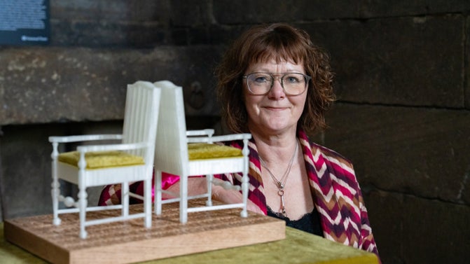 A white female with short reddish-brown wavy hair and glasses sitting next to a plinth with a model of two white chairs back to back.