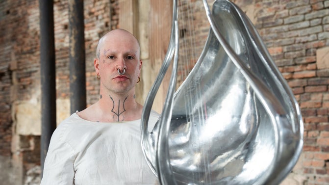 An artist wearing a white linen smock stands behind a tall silver metal sculpture in the foreground. It has a curved teardrop shape with strings attached.