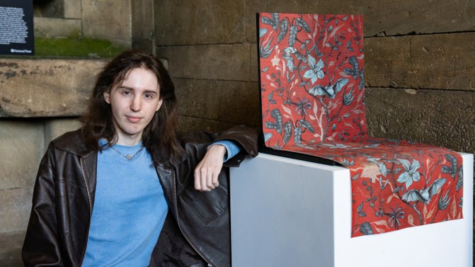 A young, white man with shoulder length brown hair sits with his arm resting on a white plinth. On top of the plinth is a sample of red wallpaper incorporating blue flowers