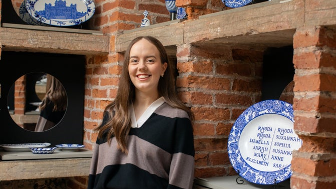 A white woman with long brown hair stands in front of a brick and wood structure with recesses. Inside each square recess is a piece of blue and white hand-painted porcelain